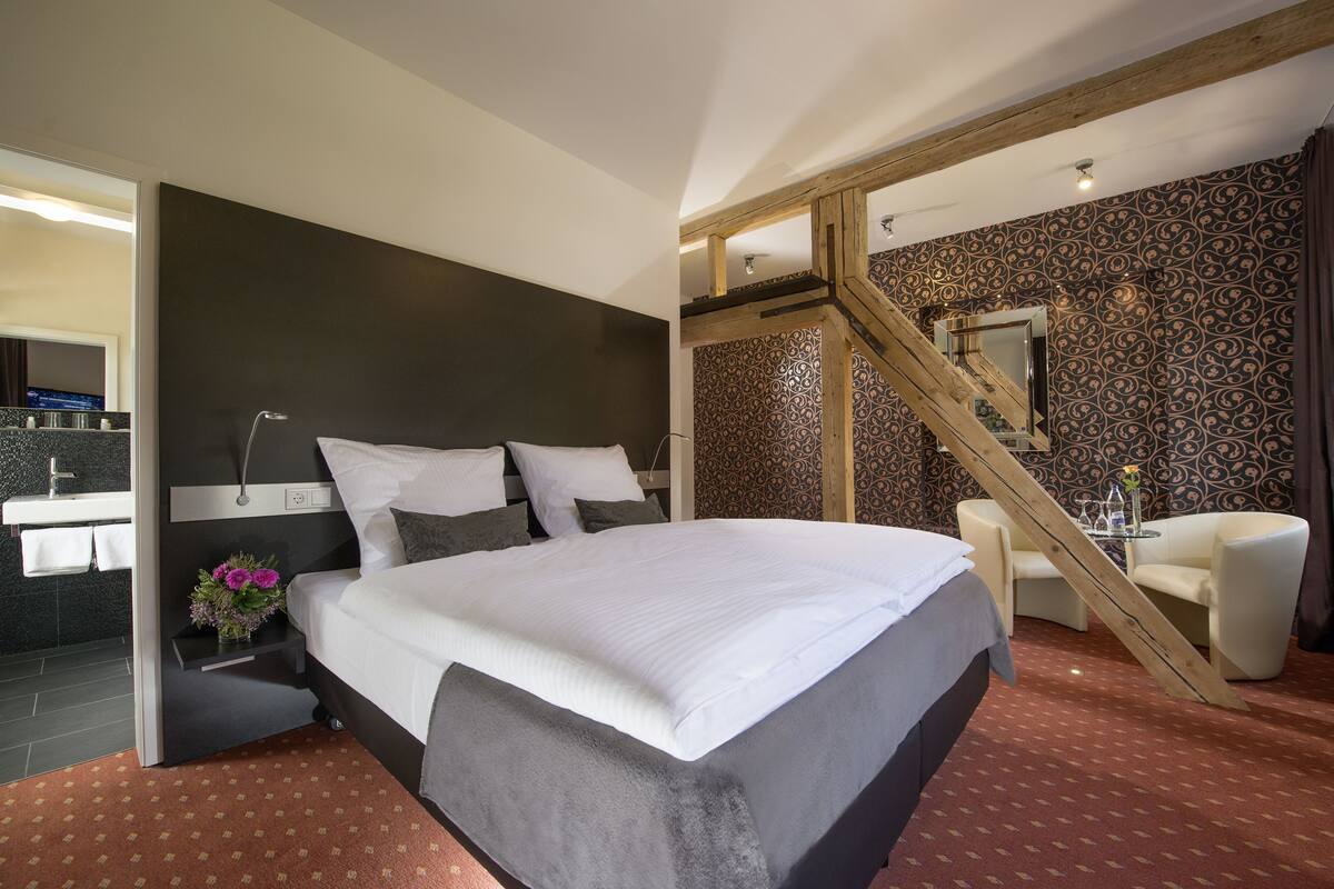 Un dormitorio moderno con una cama grande, una mesita de noche con un jarrón de flores, una silla y una escalera que conduce a un nivel superior.