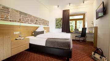 A hotel room with a bed, a desk, a chair, a television, and a stone wall.