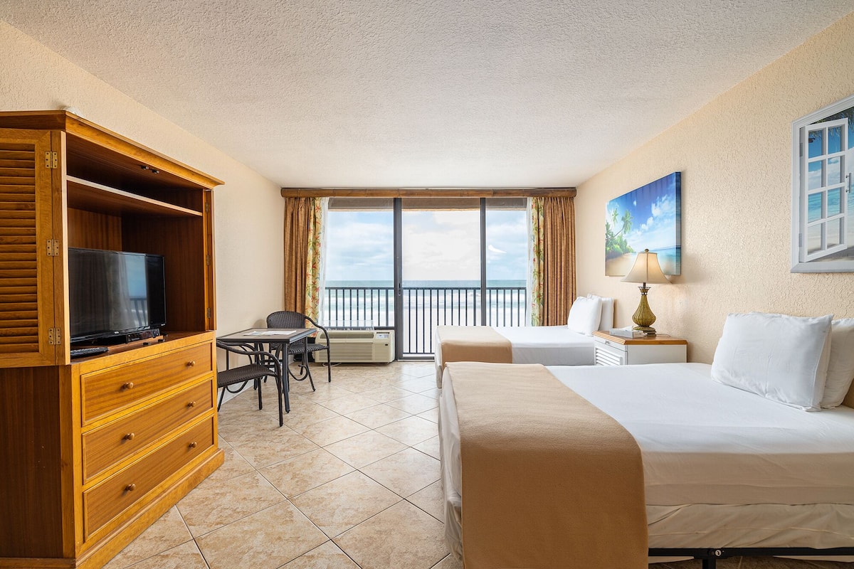 A hotel room with a TV, a desk, two beds, a balcony with a view of the ocean, and a painting on the wall.