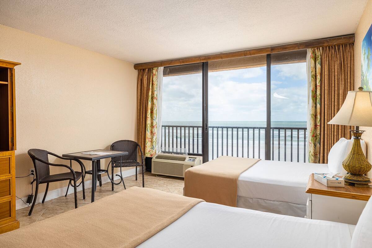 A hotel room with two beds, a desk with a chair, a window with a view of the ocean, and a balcony with a railing.