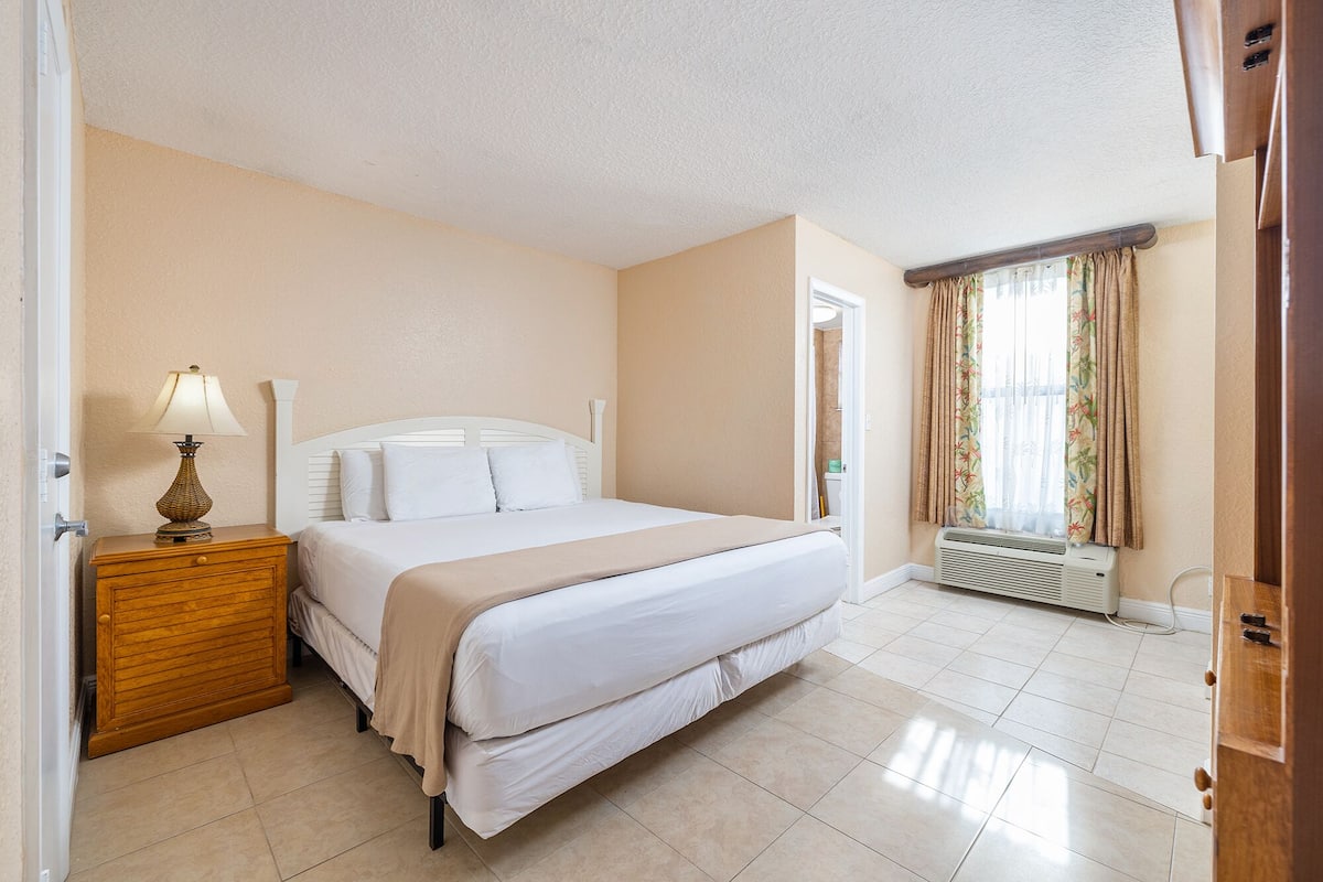 A hotel room with a bed, bedside table, lamp, air conditioning unit, and a view of the outside through a window with curtains.
