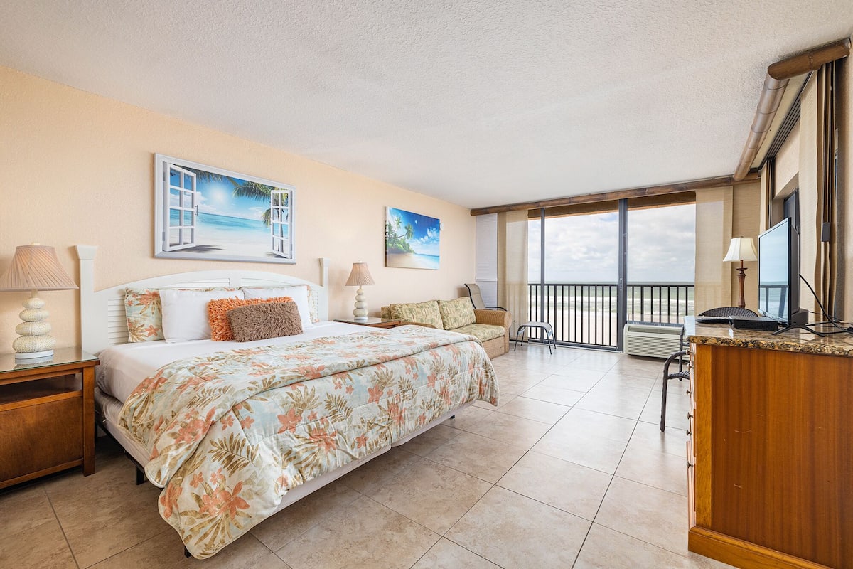 A hotel room with a large bed, a desk with a computer, a chair, and a balcony with a view of the beach.