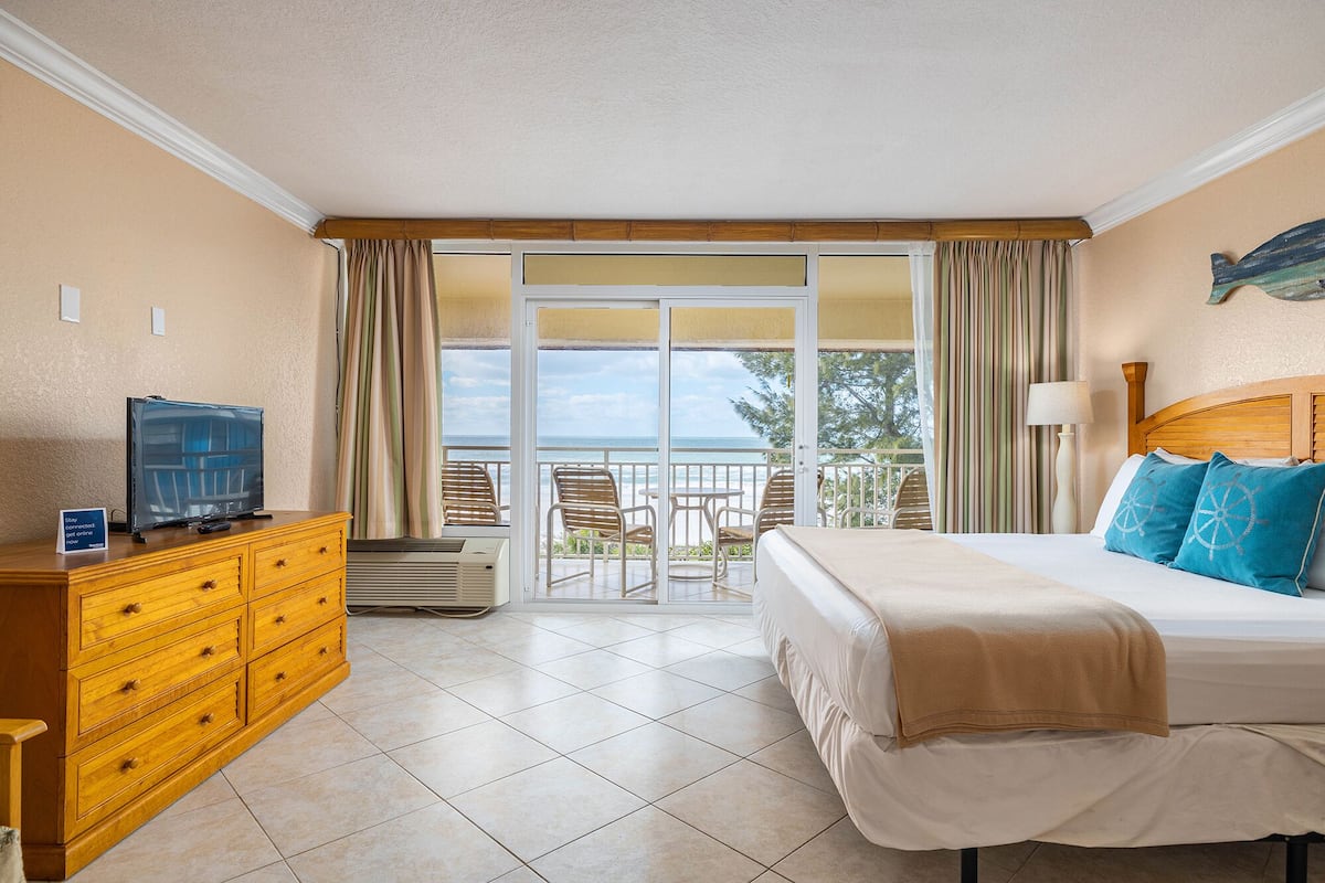 A hotel room with a bed, a dresser, a television, and a balcony with a view of the ocean.