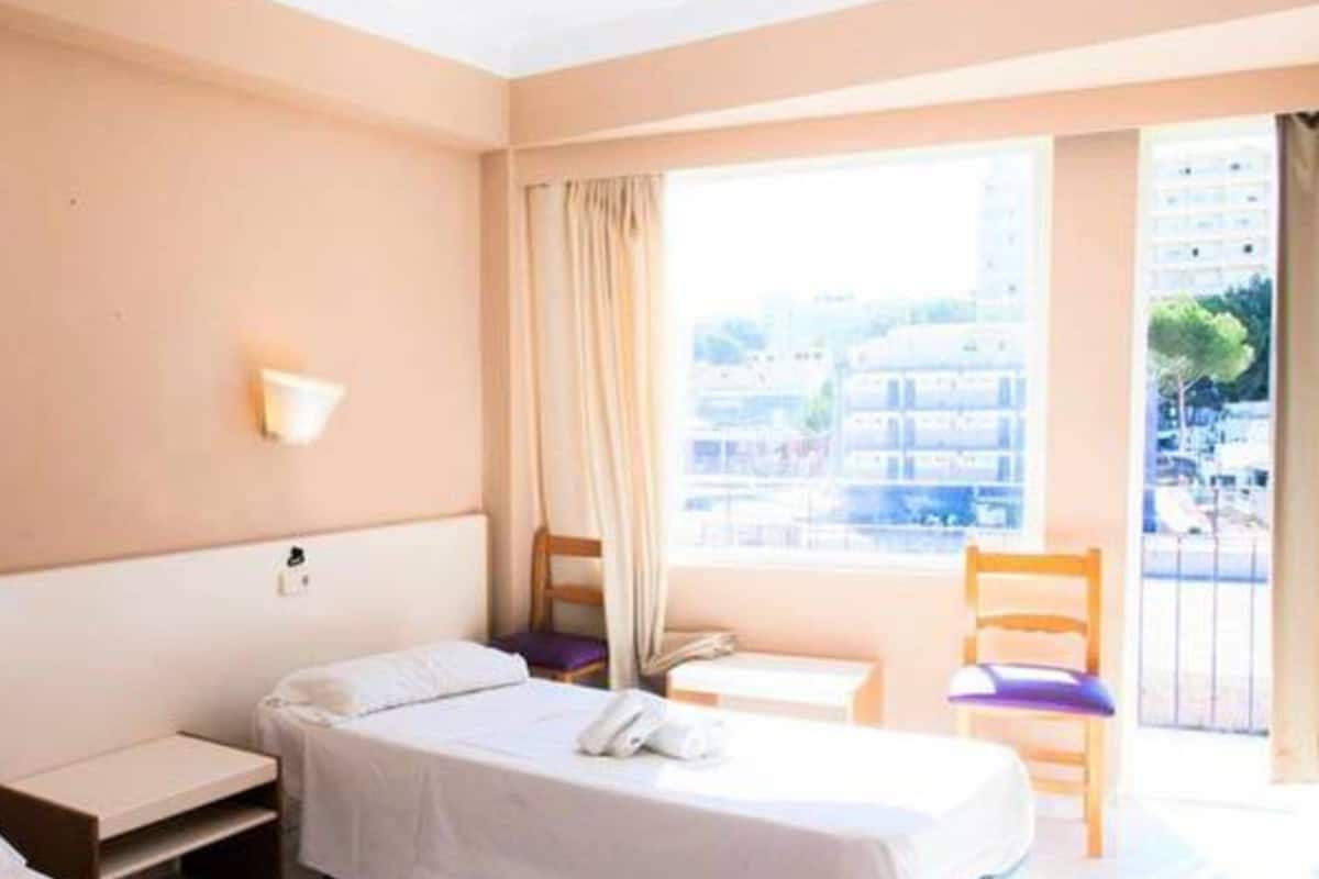 Habitación de hotel con dos camas, una mesita, una silla y una ventana con vistas a edificios.