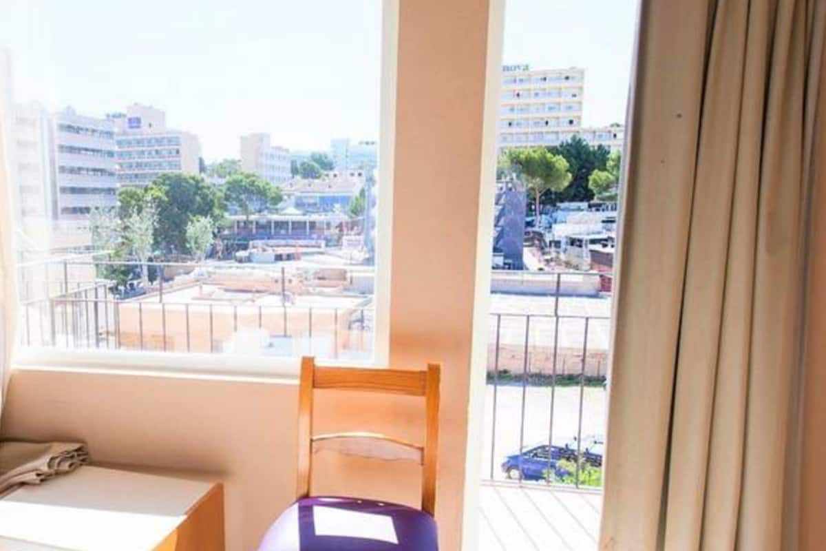 Una habitación con vista a un paisaje urbano a través de una ventana, una silla de madera y una mesita.