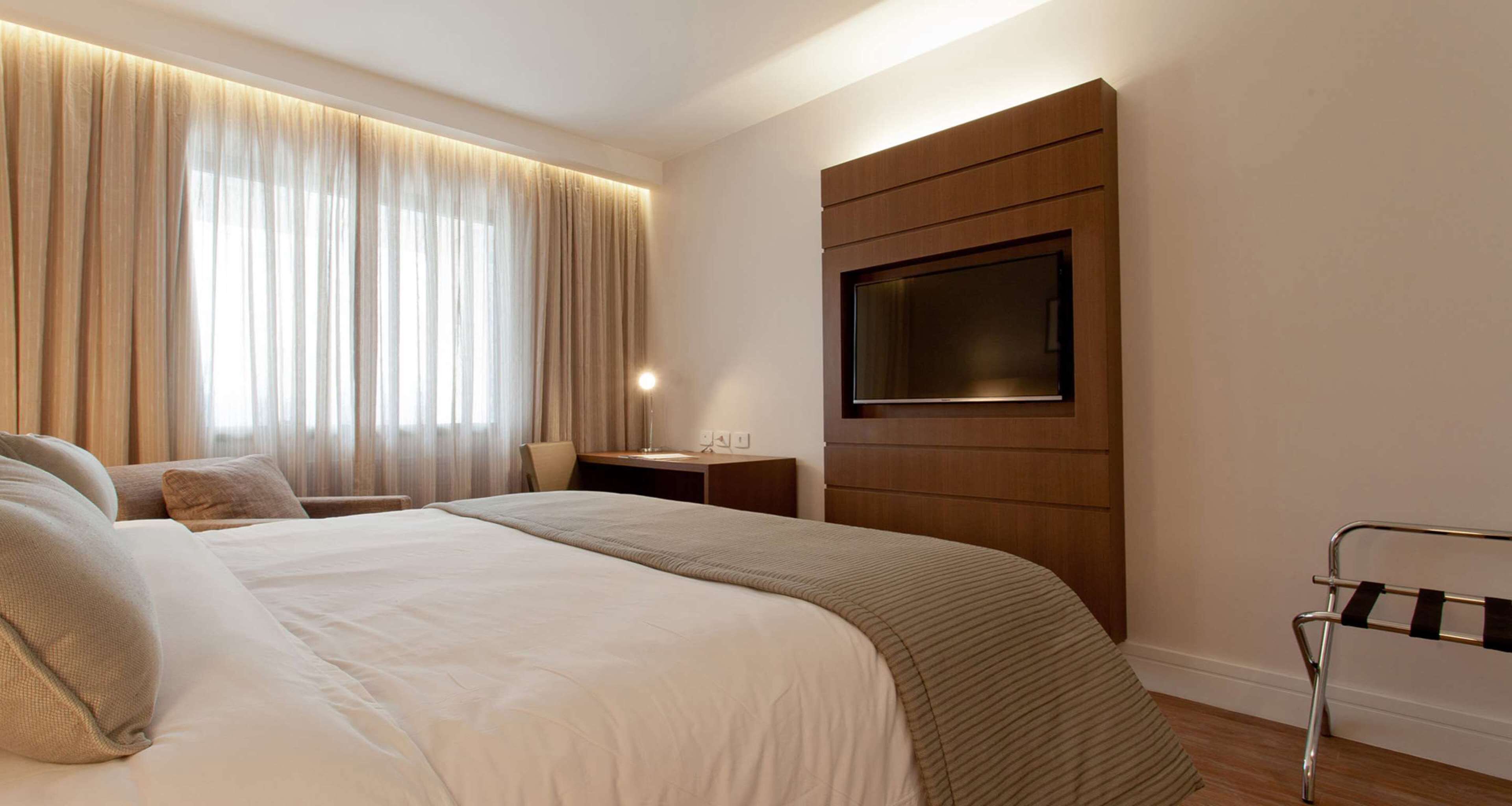 Une chambre d’hôtel avec un grand lit, un téléviseur fixé sur un panneau en bois, un bureau avec une chaise et une fenêtre avec des rideaux légers.