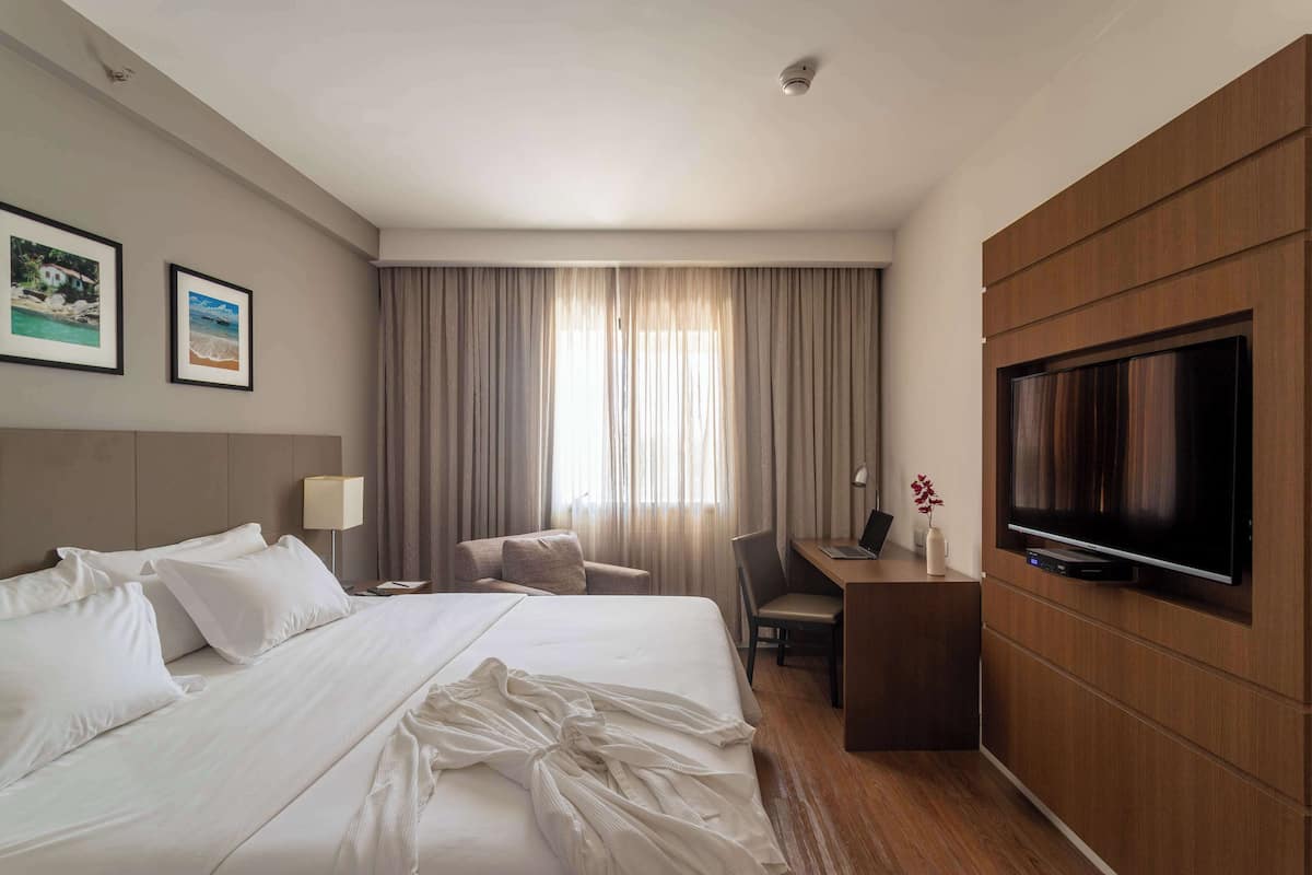 A hotel room with a large bed, a desk with a laptop, a TV, and two framed pictures on the wall.