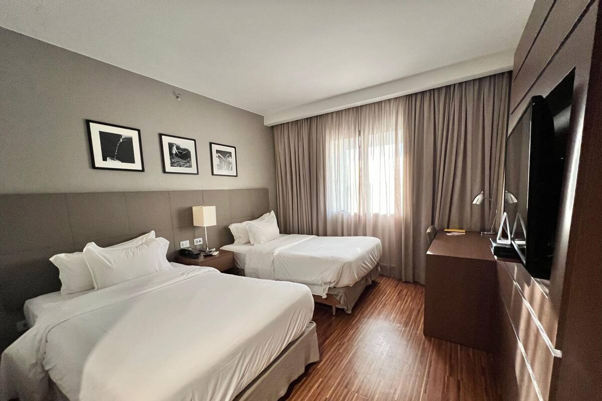 A hotel room with two beds, a desk, a TV, and a window with curtains.