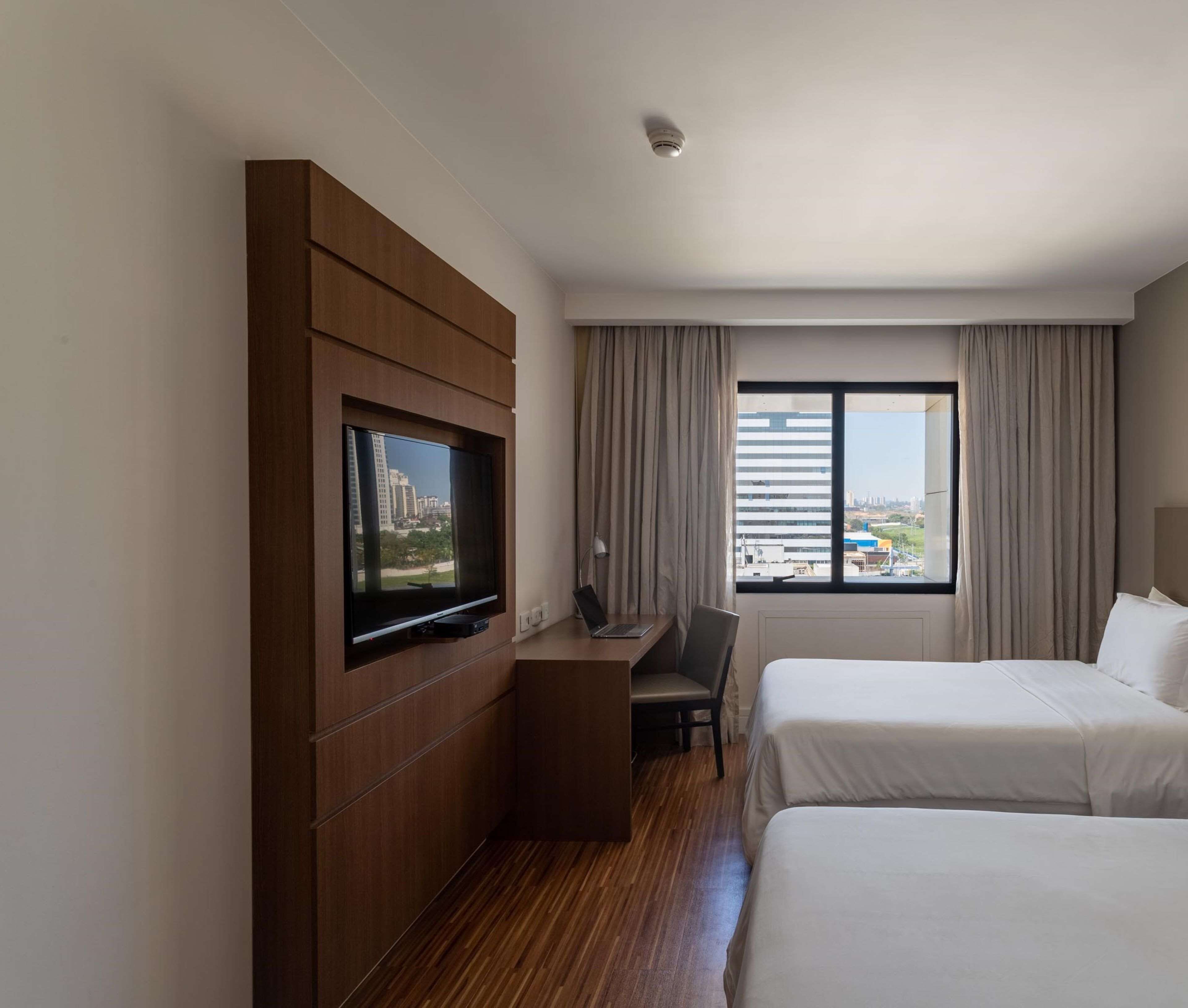 Ein Hotelzimmer mit Fernseher an einer Holzverkleidung, einem Schreibtisch mit einem Laptop, zwei Betten und einem großen Fenster mit Blick auf die Stadt.