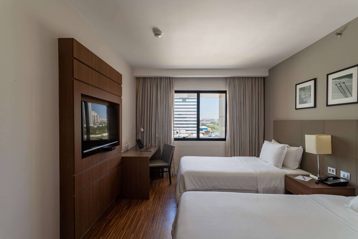 A hotel room with two beds, a desk with a laptop, a television, a window with curtains, and a city view.