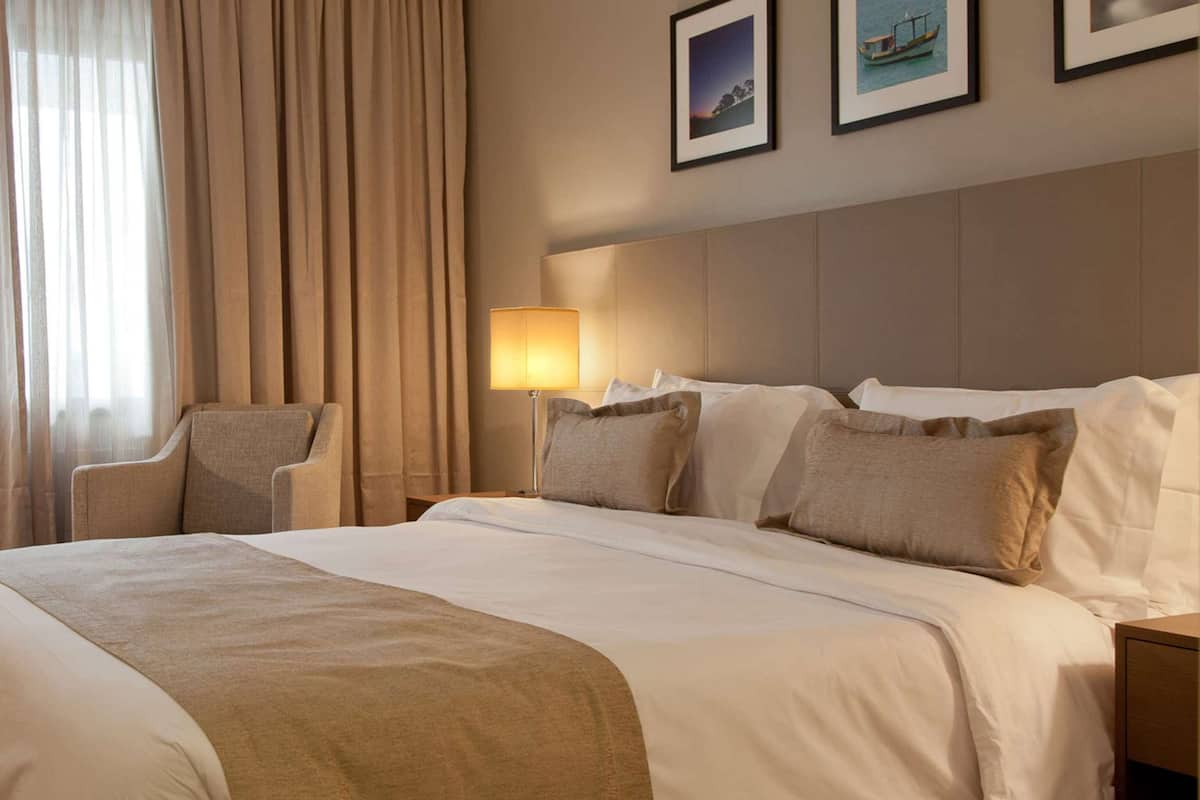 A hotel room with a large bed, a chair, a nightstand with a lamp, and curtains.