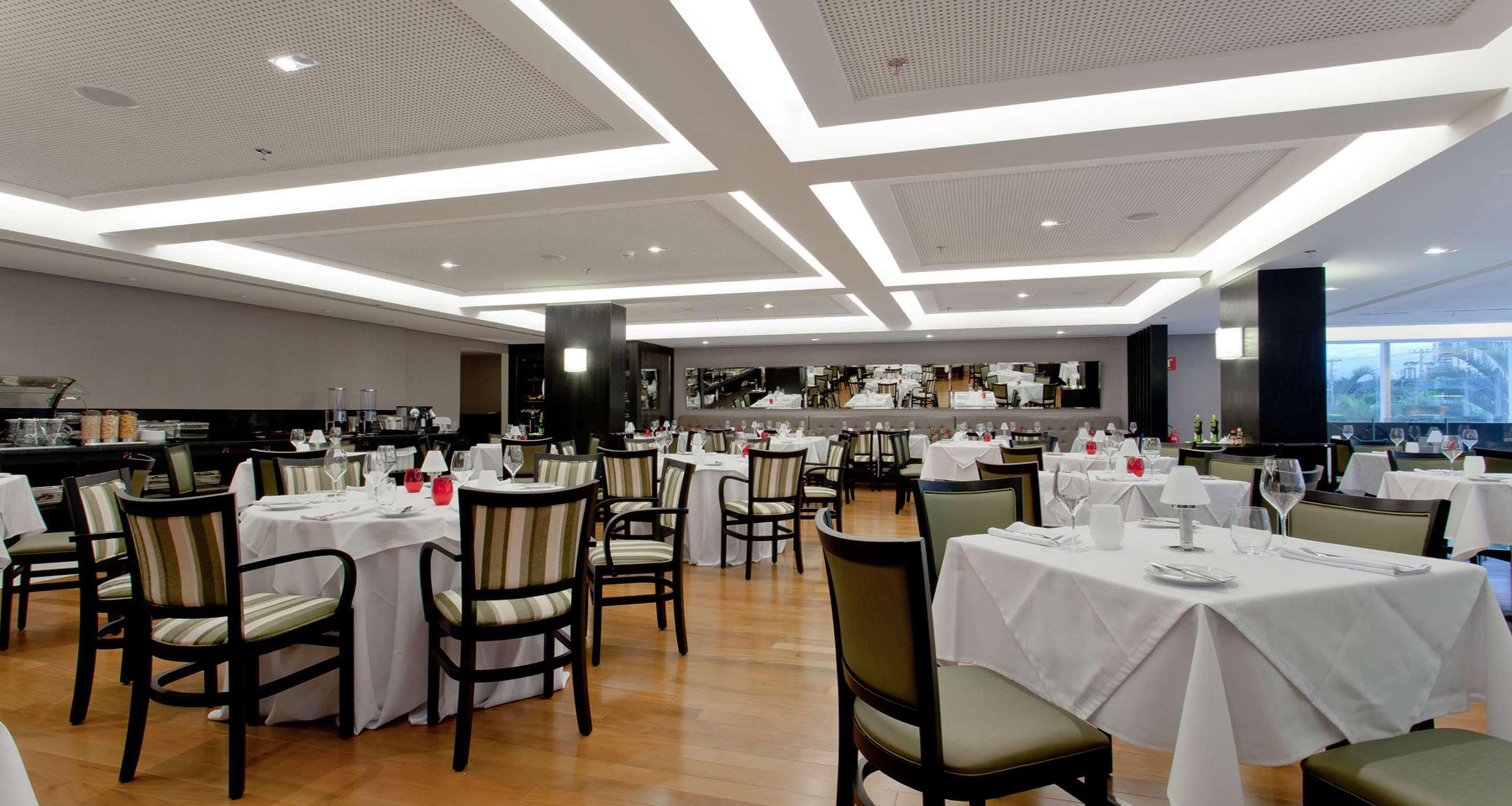 A spacious dining area with multiple tables and chairs, a kitchen in the background, and a modern ceiling design.