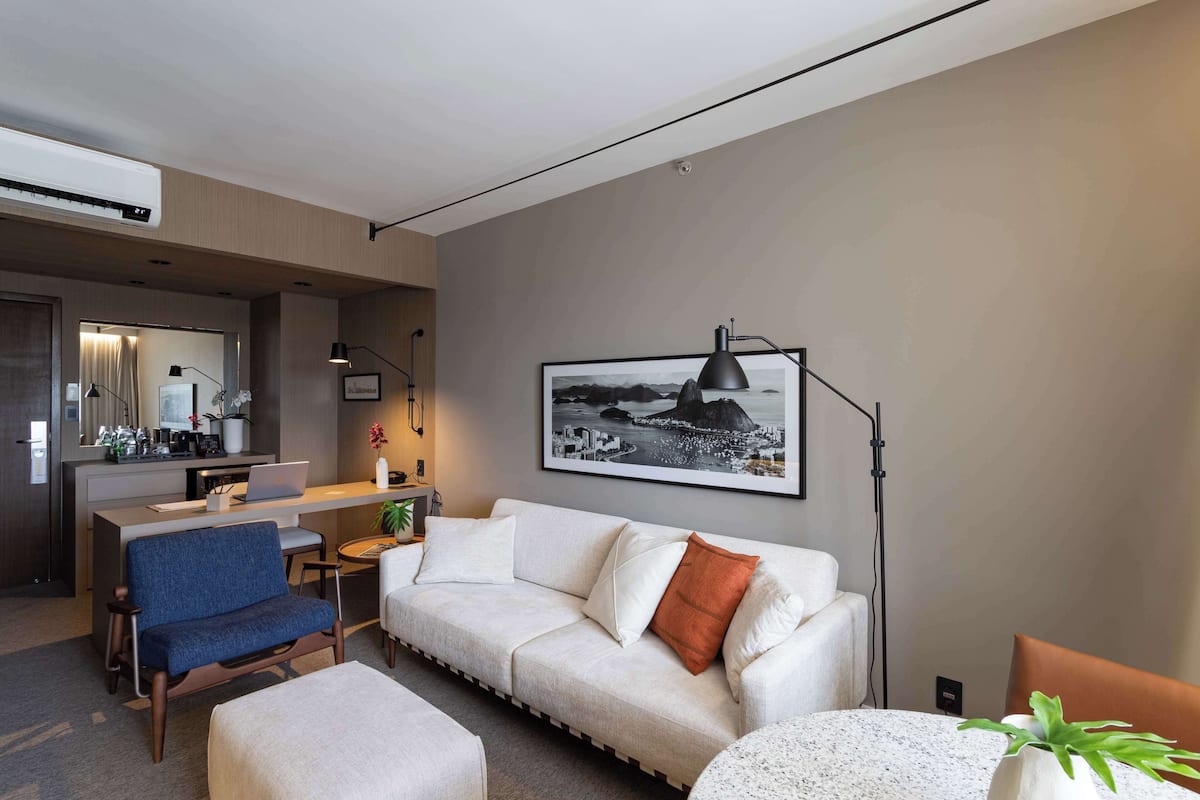A modern hotel room with a sofa, a desk, a chair, a dining table, and a large framed picture on the wall.