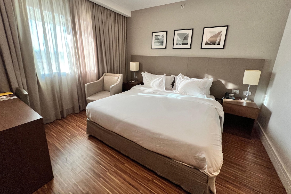 A hotel room with a large bed, a chair, a desk, and a nightstand with a lamp. There are three framed pictures on the wall.
