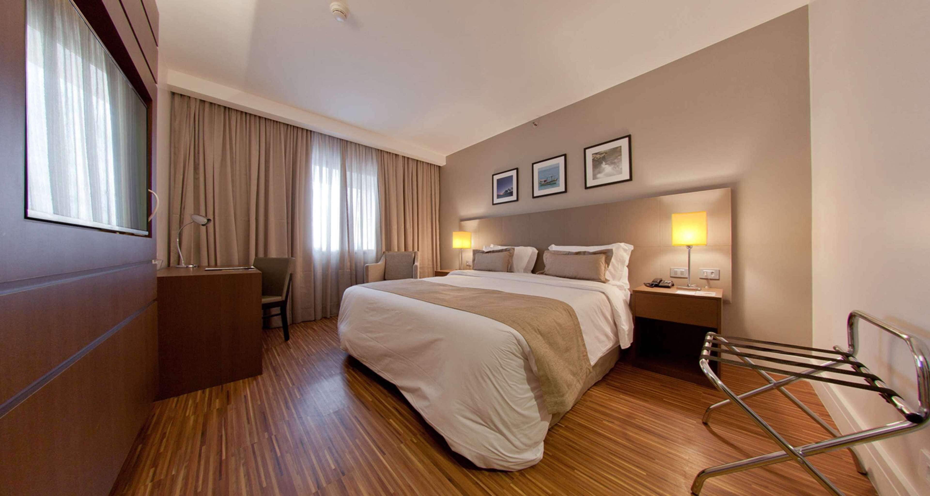 Habitación de hotel con una cama grande, un escritorio, una silla y un armario. Hay dos mesitas de noche con lámparas y una ventana con cortinas.