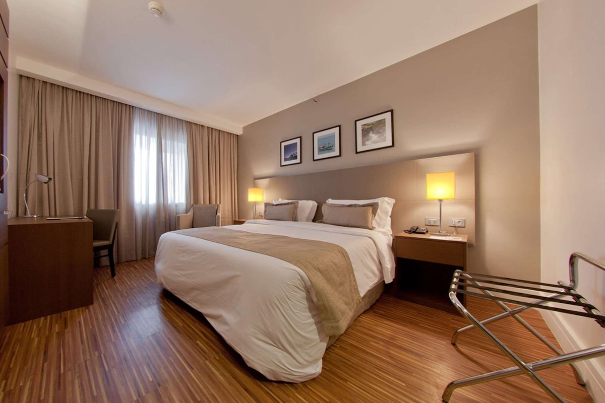 A hotel room with a large bed, a desk, a chair, and a wardrobe. There are two bedside tables with lamps and a window with curtains.