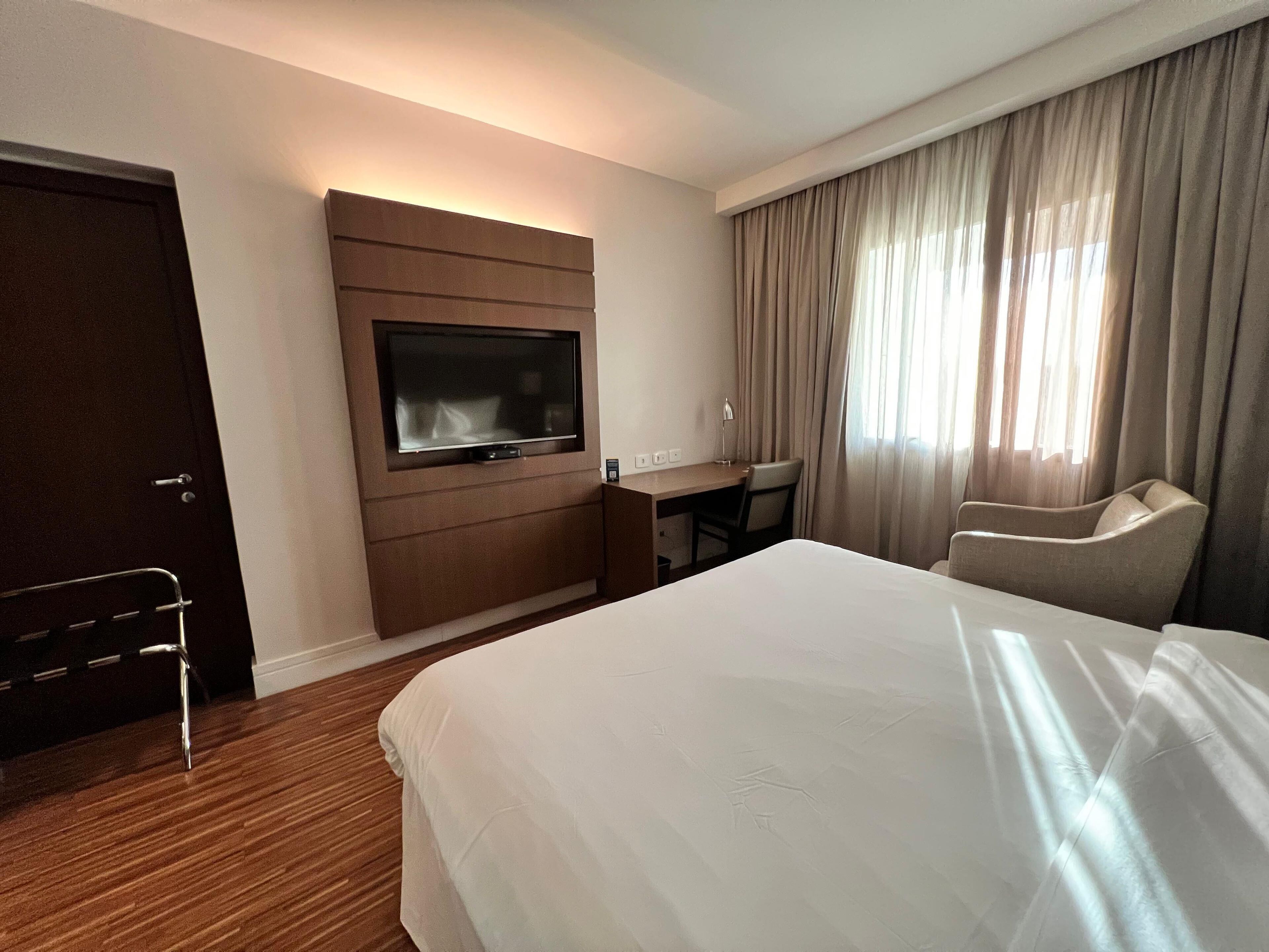 A hotel room with a large bed, a desk with a chair, a TV mounted on the wall, and a door.