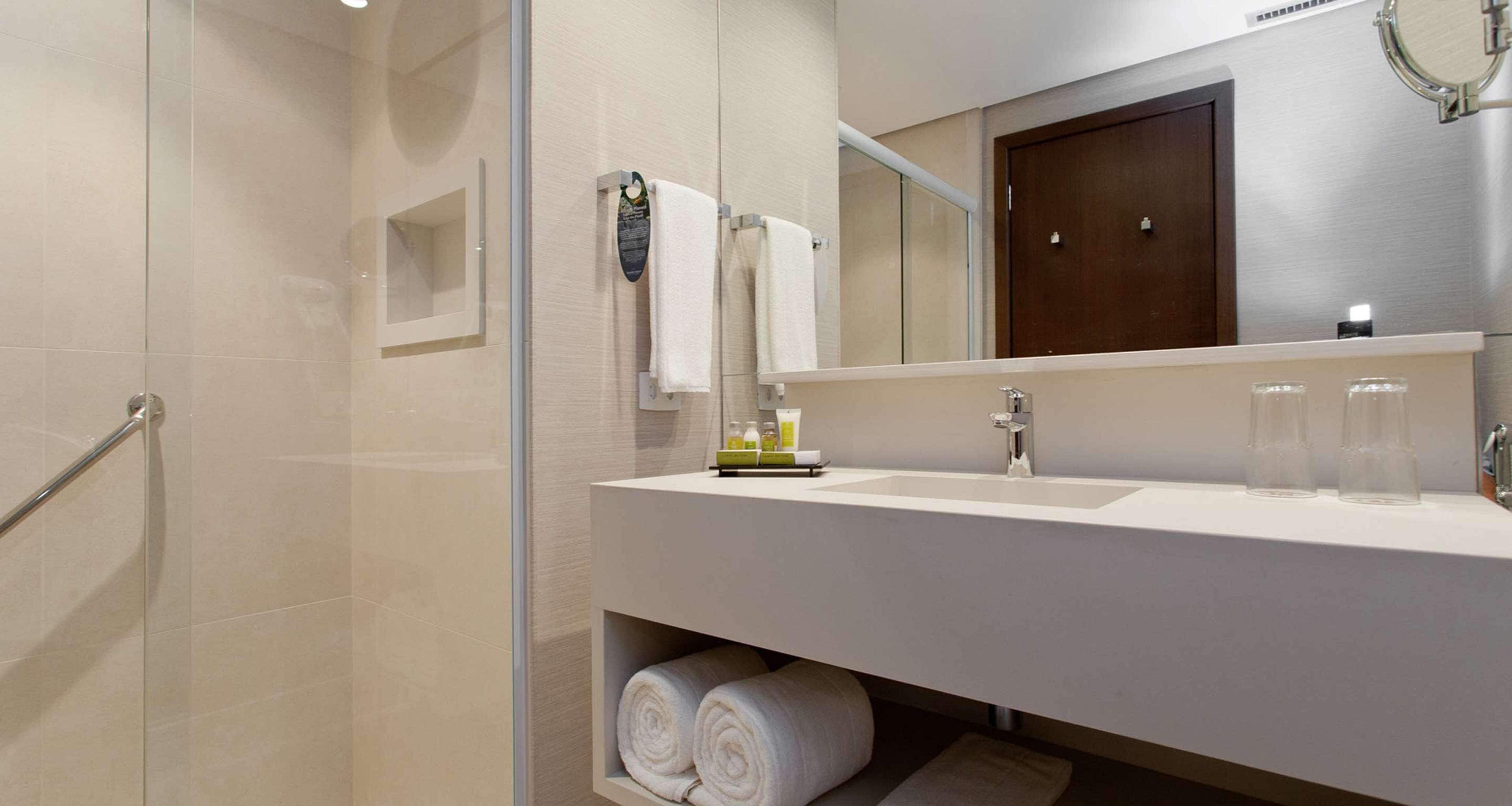 A modern bathroom with a glass shower enclosure, a white sink with a mirror above, and neatly rolled towels on a shelf.