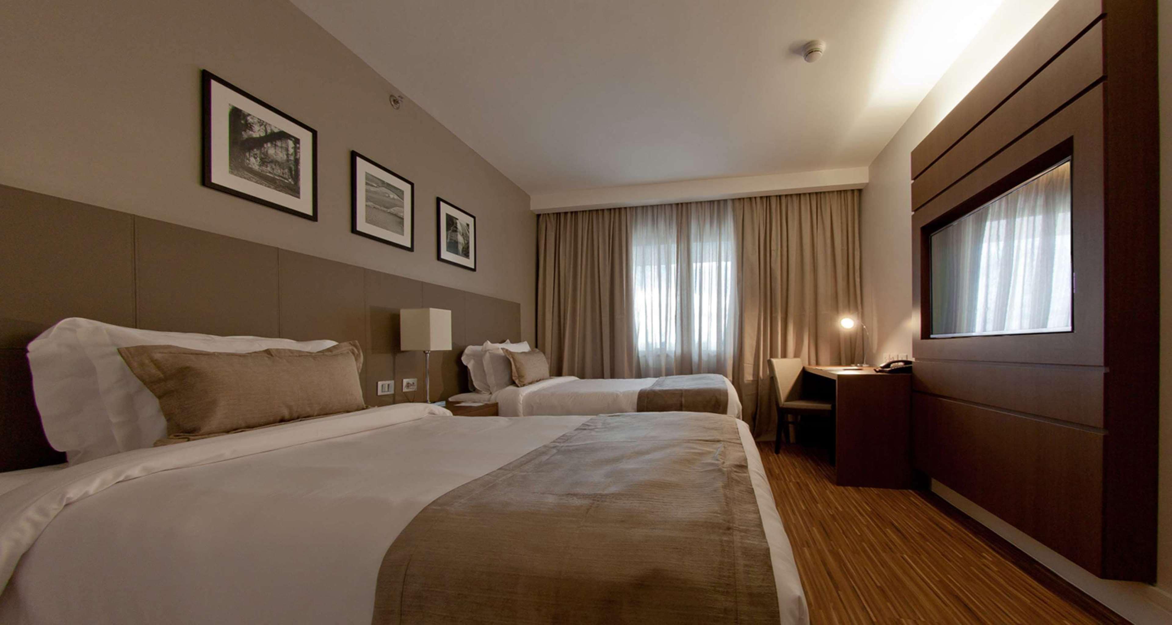 A hotel room with two beds, a desk, a chair, and a large window with curtains.