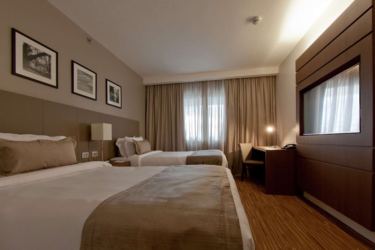 A hotel room with two beds, a desk, a chair, and a large window with curtains.