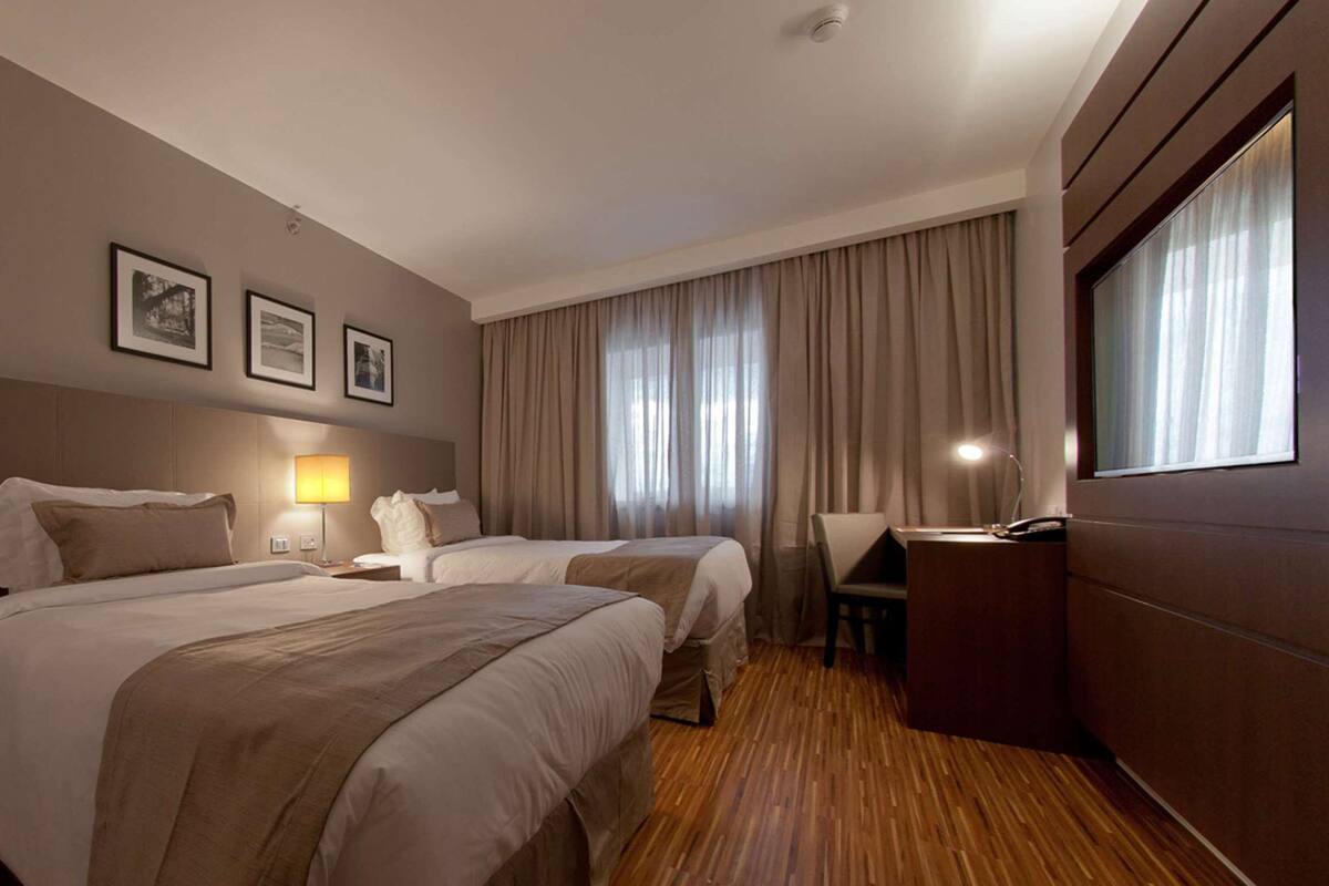 A hotel room with two beds, a desk, a chair, a lamp, and a large window with curtains.