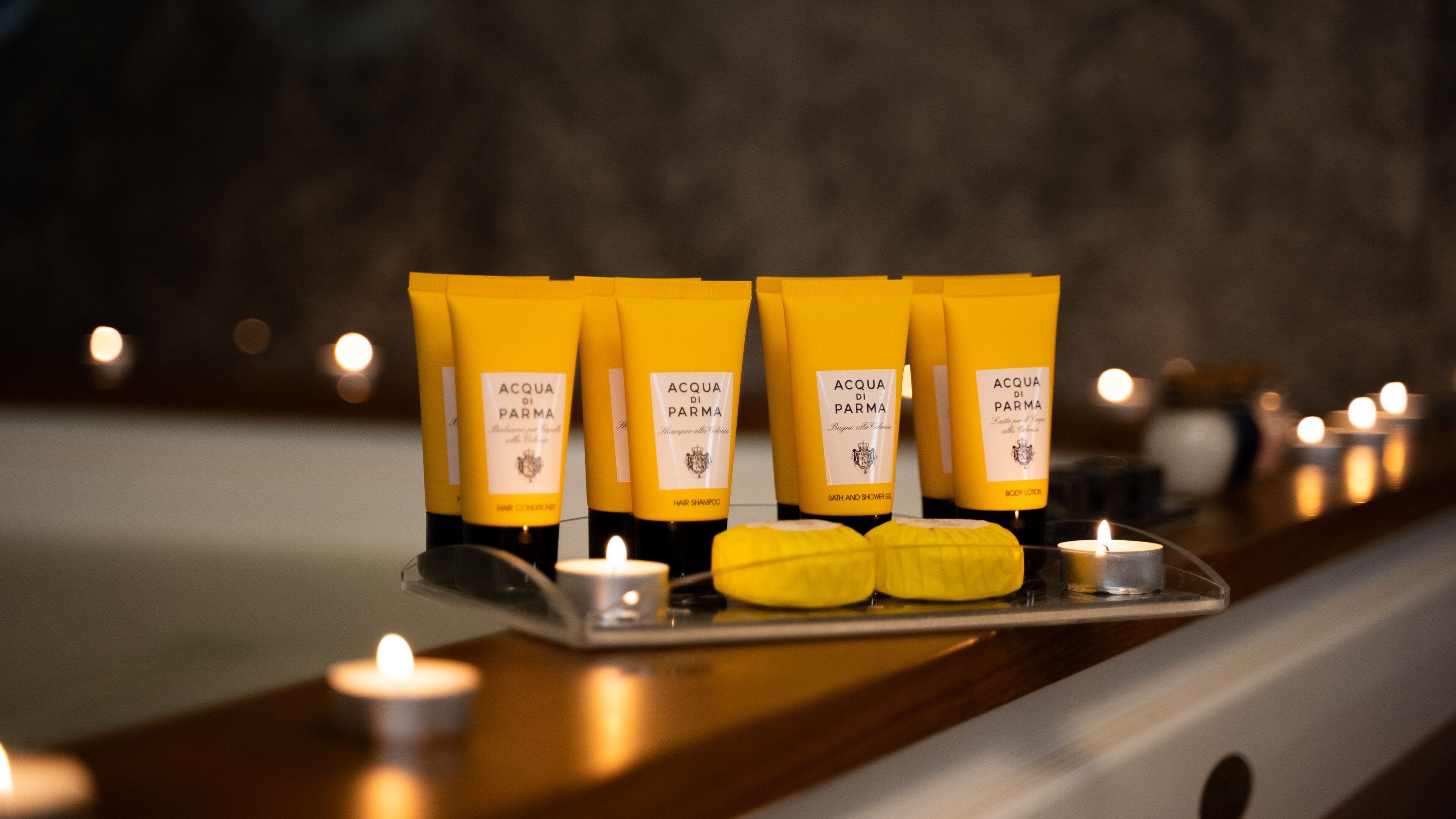 A tray with four yellow tubes labeled "ACQUA DI PARMA" on a wooden surface, accompanied by candles.