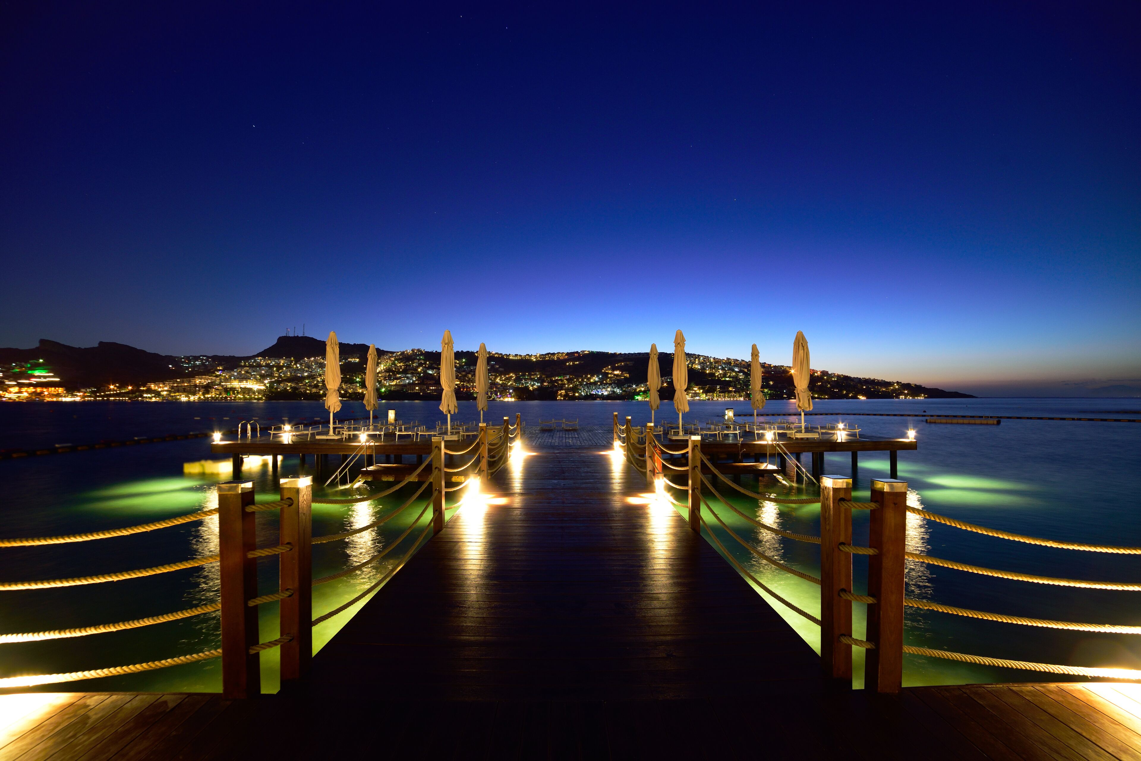 A wooden pier with rope railings leading to a dock, overlooking a calm body of water and a cityscape at night.
