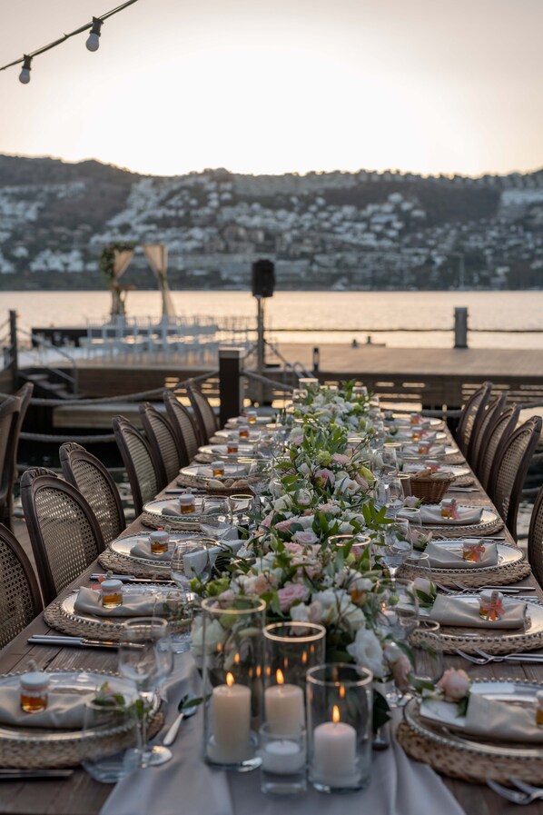 A long dining table set for a meal with candles, glasses, and plates, overlooking a body of water and snow-covered hills. - Mivara Luxury Resort & Spa Bodrum (Bodrum)