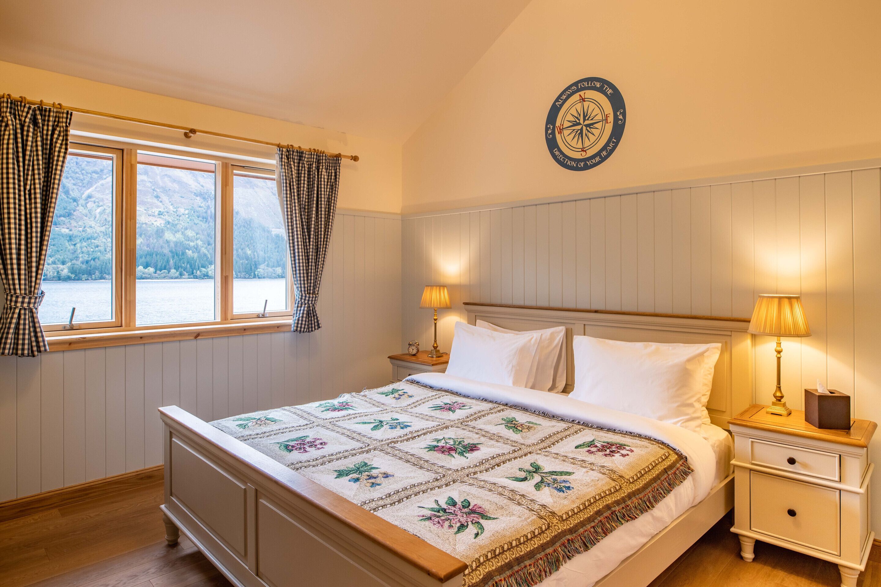 A bedroom with a bed, two bedside lamps, a nightstand, a window with curtains, and a wall-mounted compass.