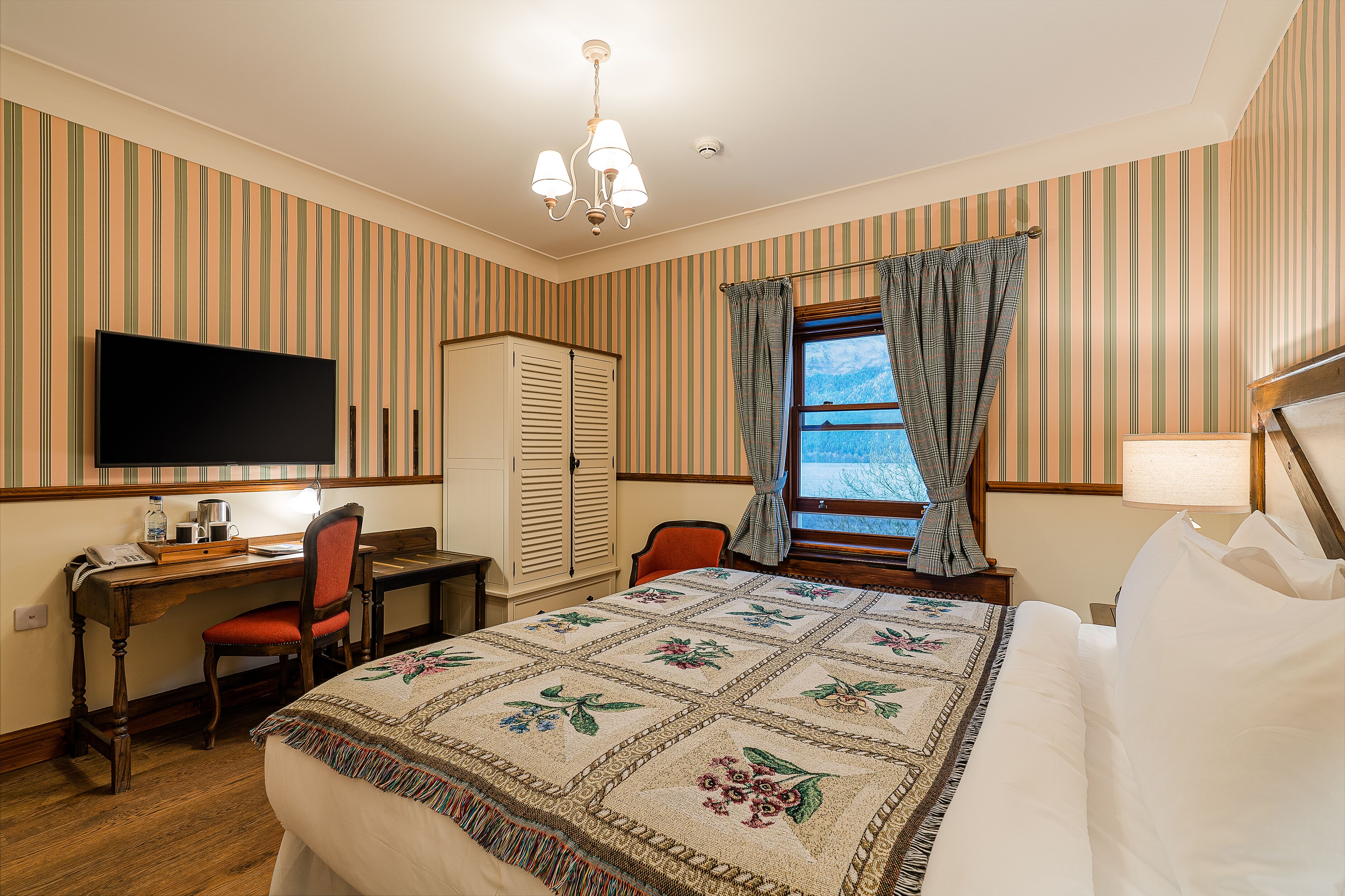A hotel room with a bed, a desk with a chair, a television, a wardrobe, and a window with curtains.