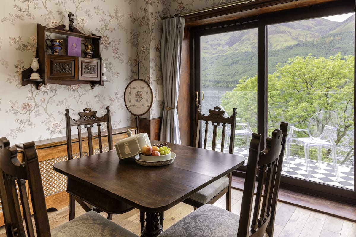 Une table à manger avec des chaises, un panier de fruits et une assiette décorative. Une fenêtre donne sur un paysage montagneux.