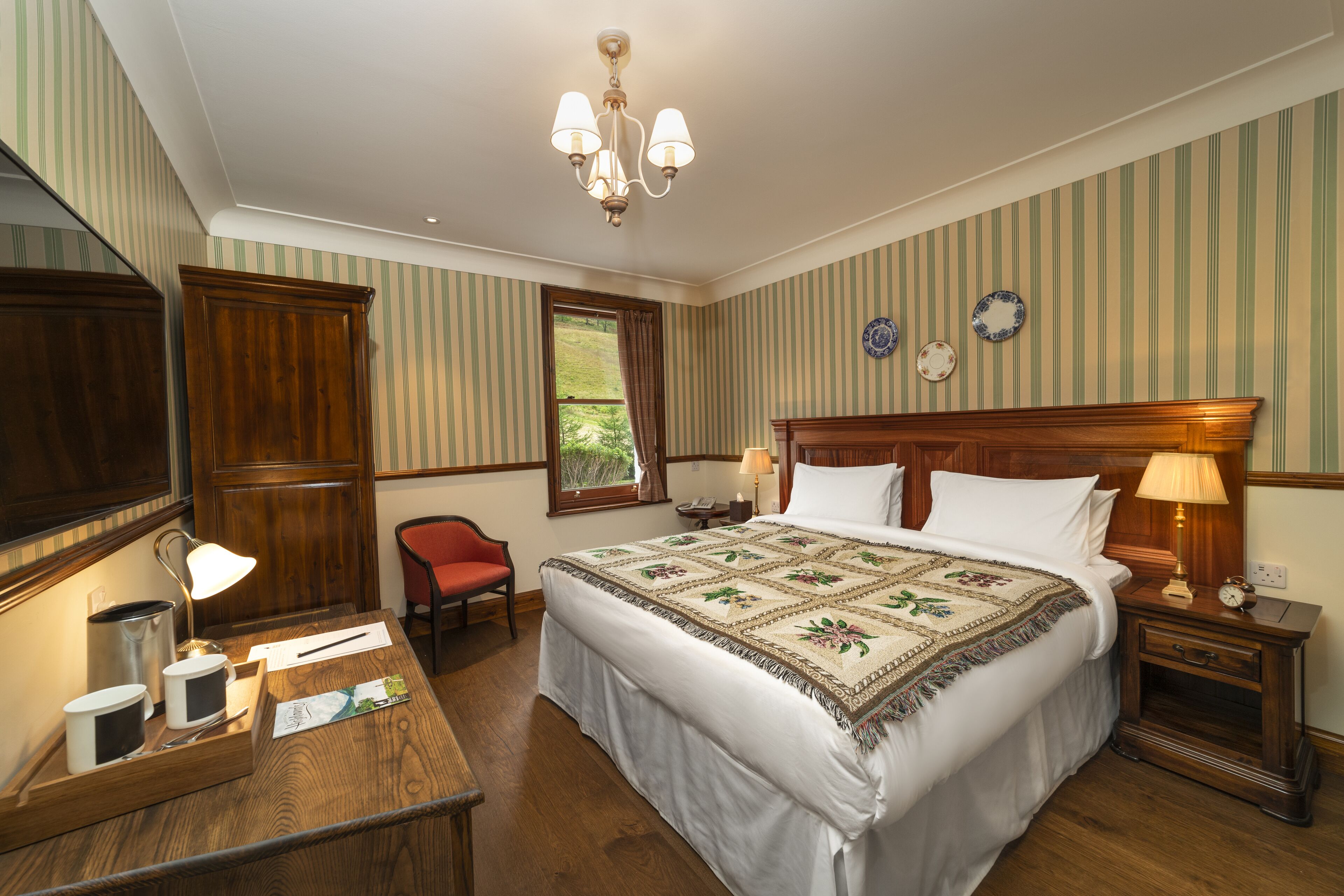 A hotel room with a large bed, wooden headboard, a desk with a lamp, a chair, and a window with curtains.