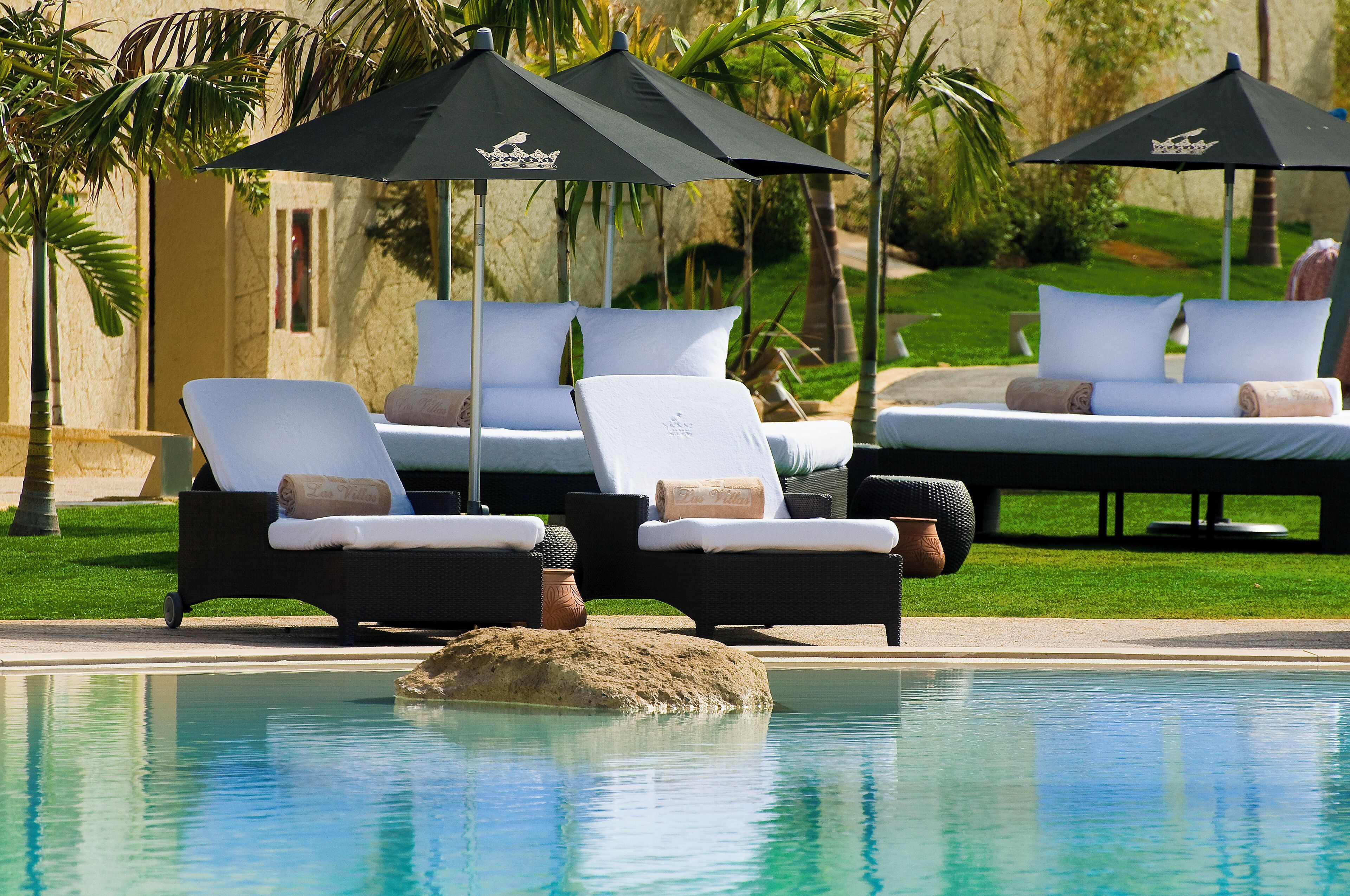 Un área junto a la piscina con sillones de descanso y sombrillas, una piscina cristalina y un jardín ajardinado.