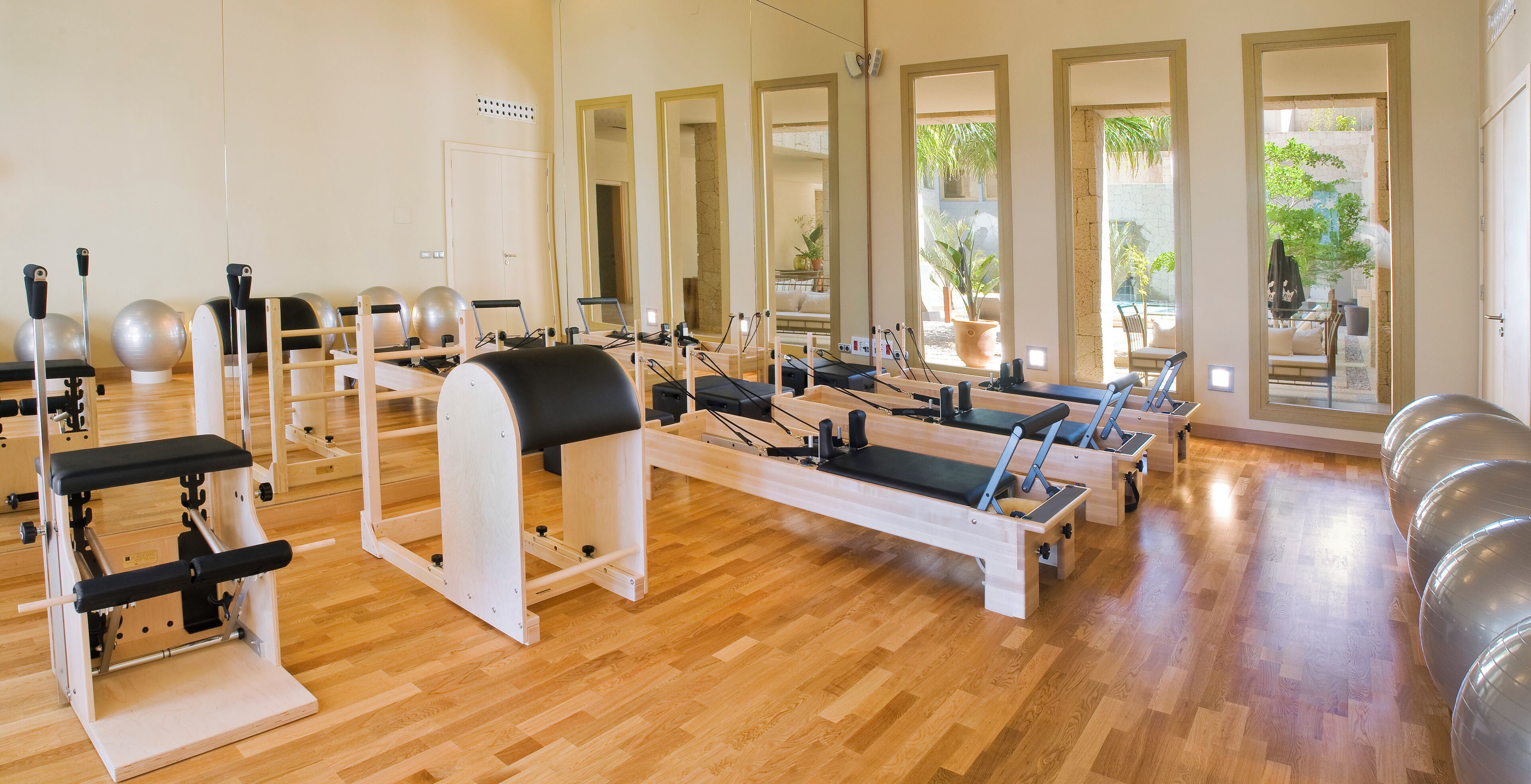 A Pilates studio with wooden floors, exercise equipment, and large windows offering views of the outdoors.
