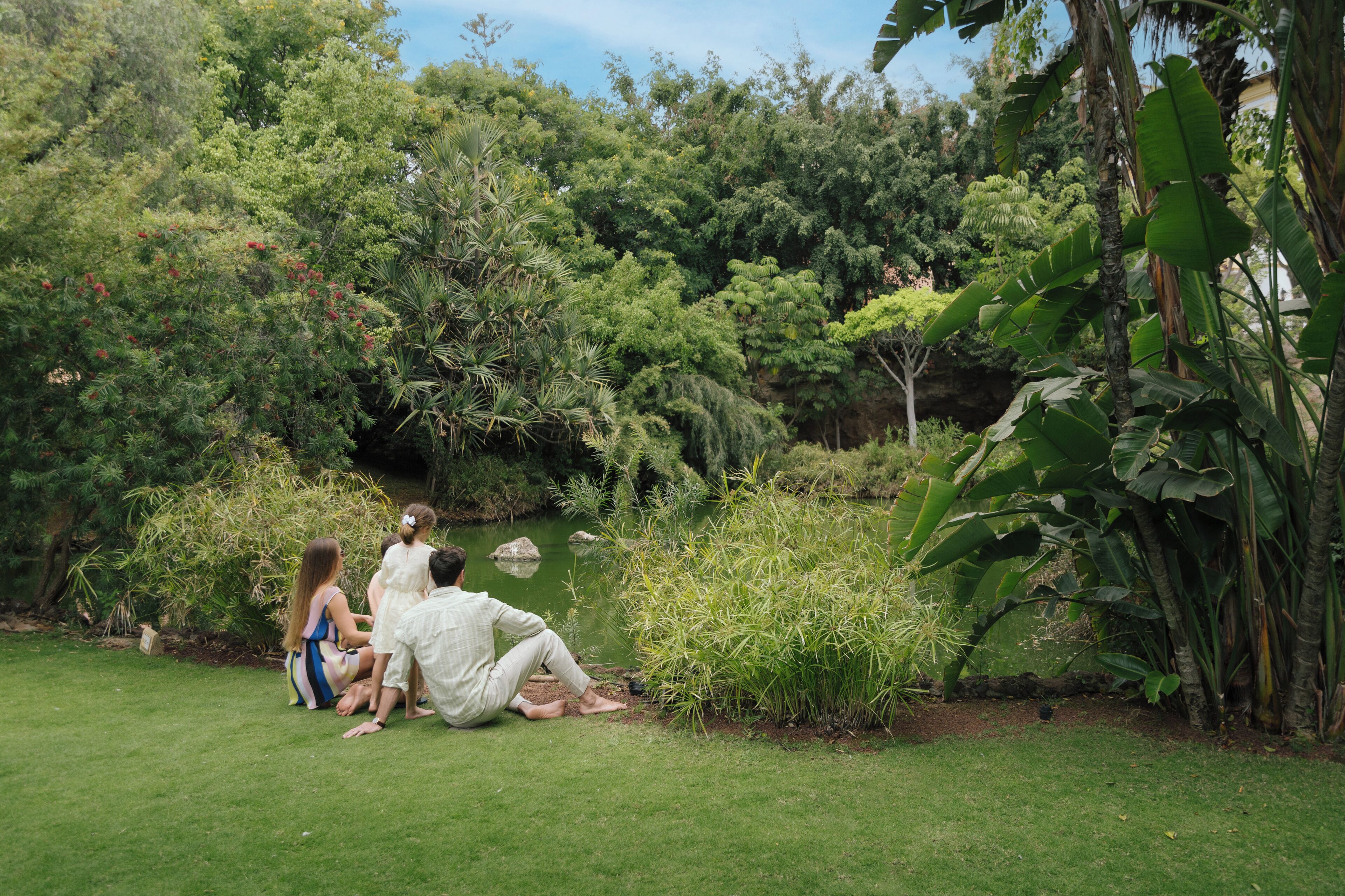 A group of people sitting on grass near a pond in a lush garden with various trees and plants.