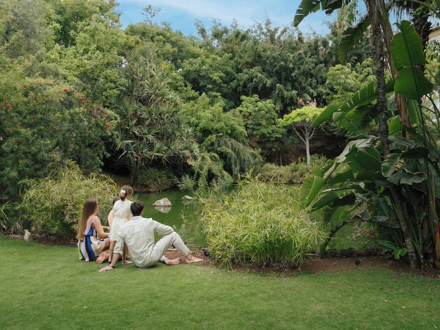 A group of people sitting on grass near a pond in a lush garden with various trees and plants.