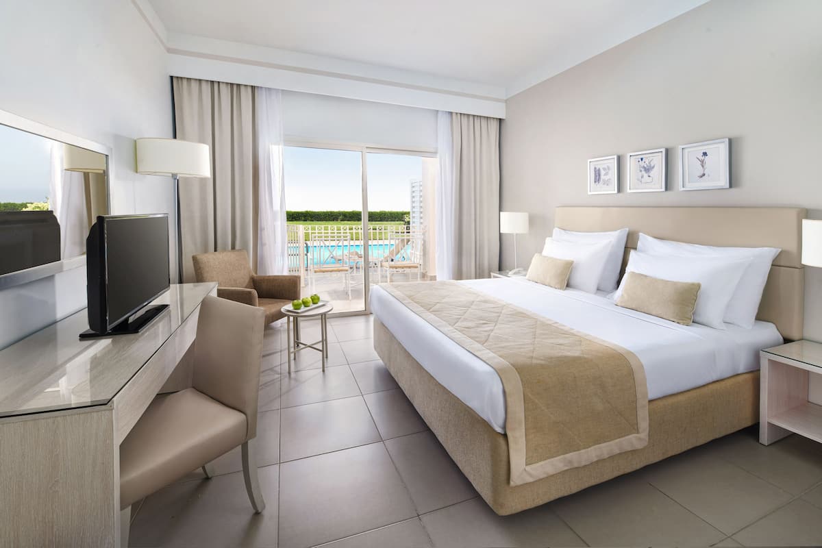 A hotel room with a bed, a desk, a chair, a TV, a small table with fruit, and a balcony with a pool.