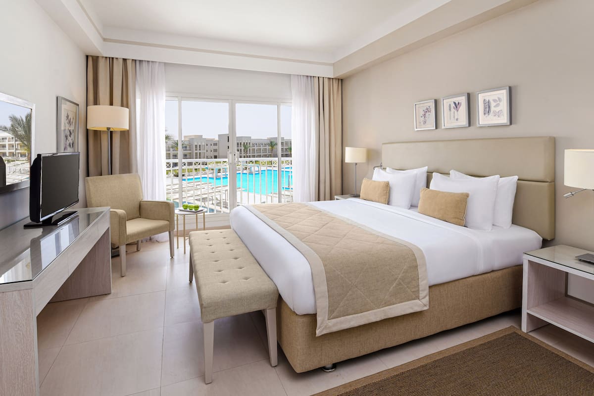 A hotel room with a bed, a desk, a chair, a TV, a small table, and a view of a swimming pool.