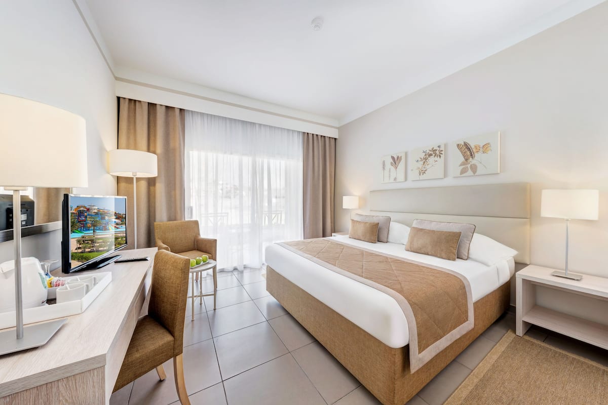 A hotel room with a bed, a desk, a chair, a TV, and a balcony with curtains.