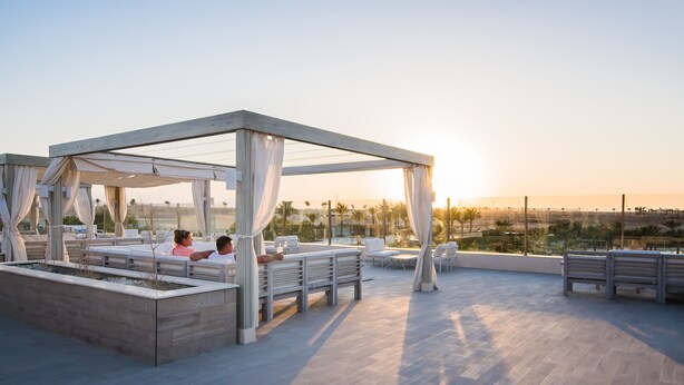 A rooftop terrace with a wooden pergola, white cushions, and a view of palm trees and buildings. - JAZ Aquaviva - All inclusive (Makadi Bay)