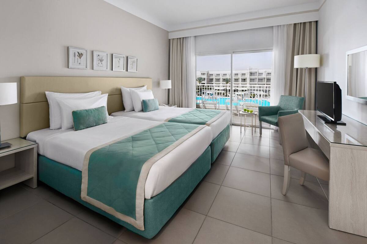 A hotel room with a large bed, a desk, a chair, a TV, and a balcony with a pool view.