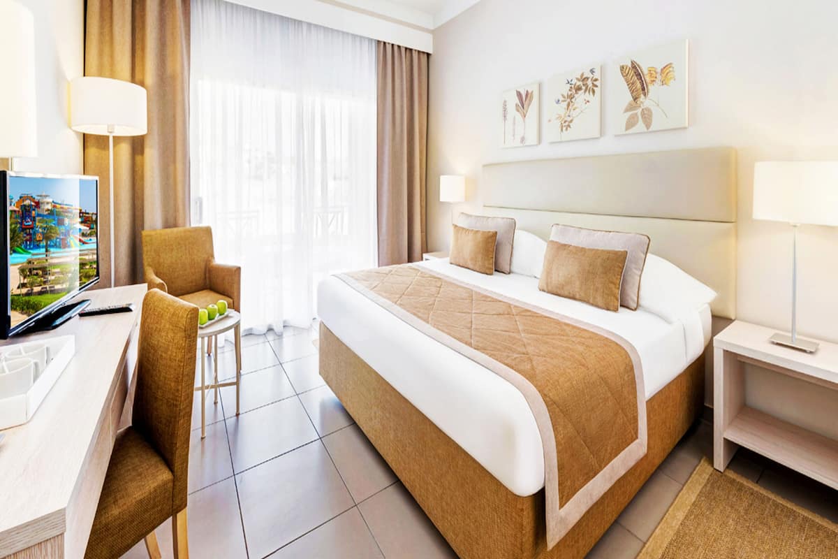 A hotel room with a large bed, a desk with a laptop, a chair, and a TV displaying a scenic view.