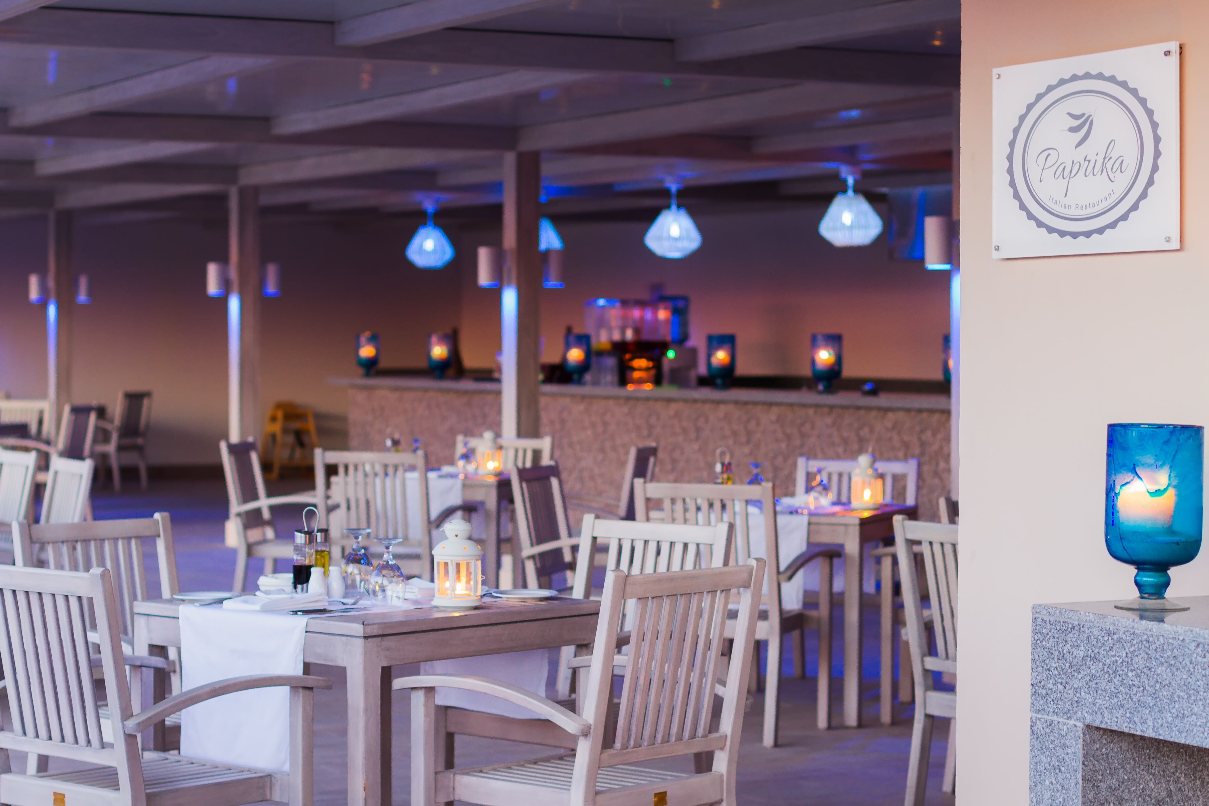 A restaurant interior with white wooden tables and chairs, hanging pendant lights, and a blue candle holder on a stone pedestal.