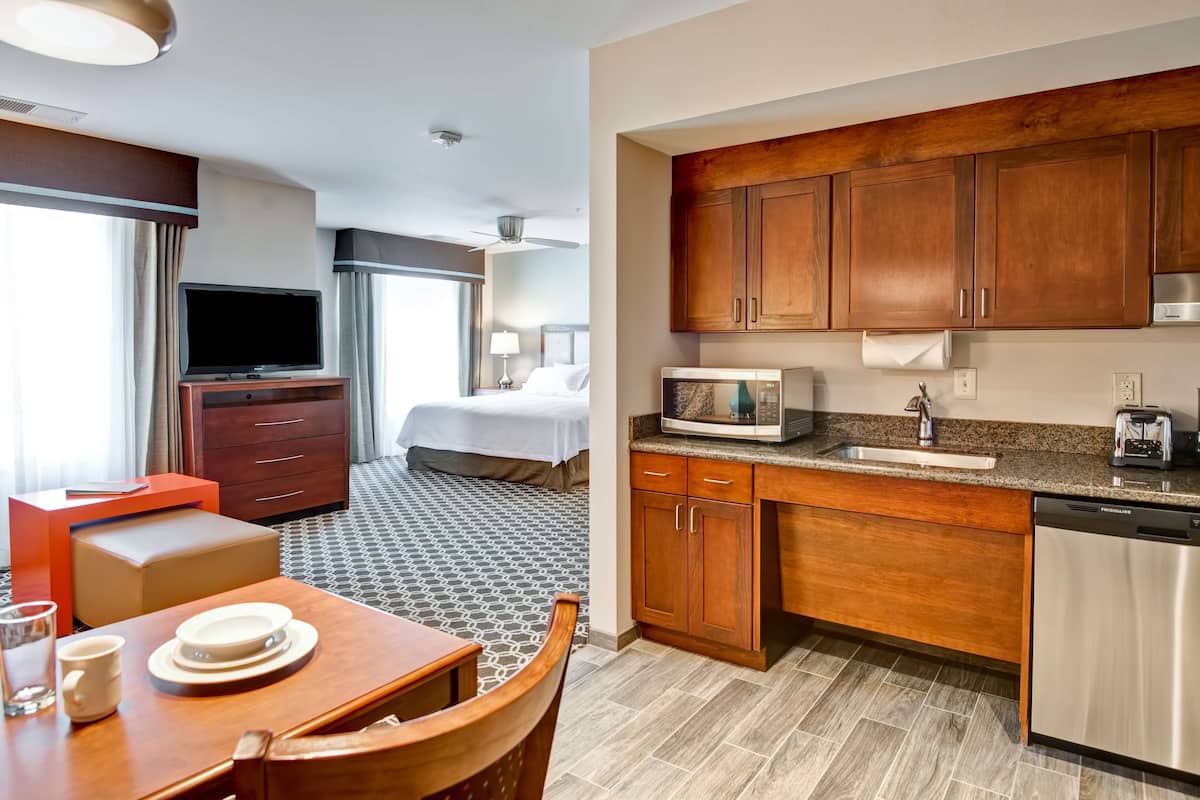 A hotel room with a bed, a TV, a dining area with a table and chairs, and a kitchenette with wooden cabinets and a microwave.