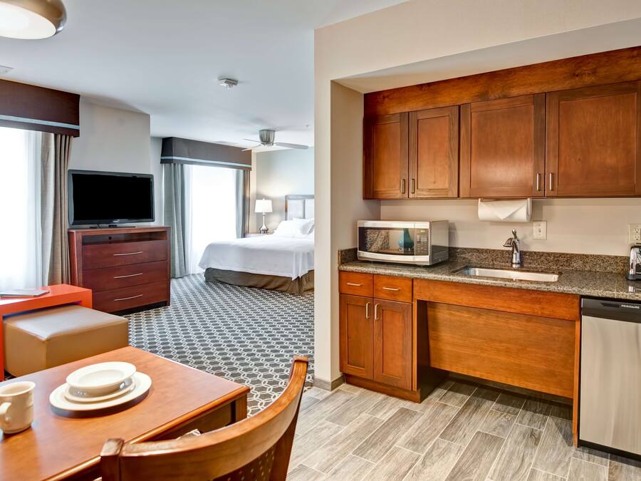 A hotel room with a bed, a TV, a dining area with a table and chairs, and a kitchenette with wooden cabinets and a microwave.