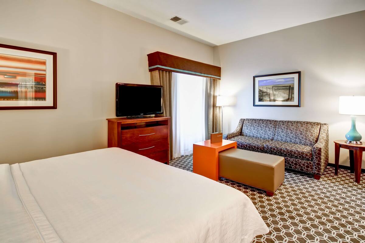 A hotel room with a large bed, a TV on a wooden stand, a patterned sofa, a small table with a lamp, and a painting on the wall.
