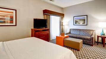 A hotel room with a large bed, a TV on a wooden stand, a patterned sofa, a small table with a lamp, and a painting on the wall.