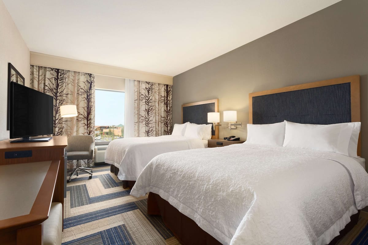 A hotel room with two beds, a desk with a television, a chair, and a view of the outdoors.