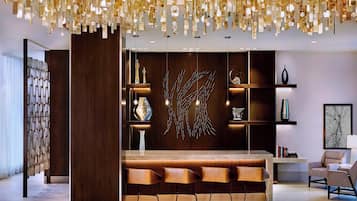 A modern reception area with a wooden reception desk, three bar stools, a decorative wall art piece, and a chandelier with geometric light fixtures.