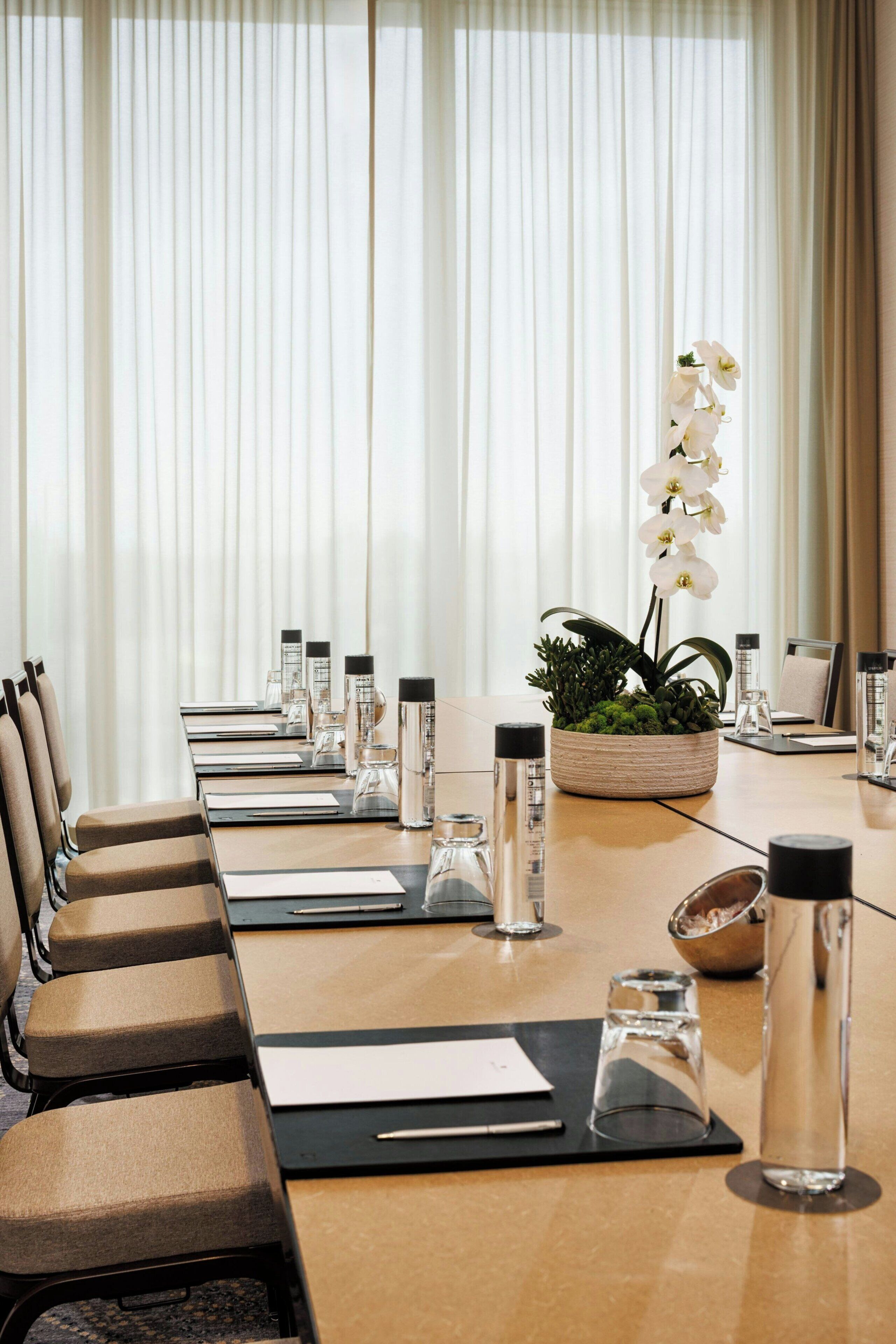 A conference room with a long table, chairs, water bottles, a potted plant, and a window with curtains.